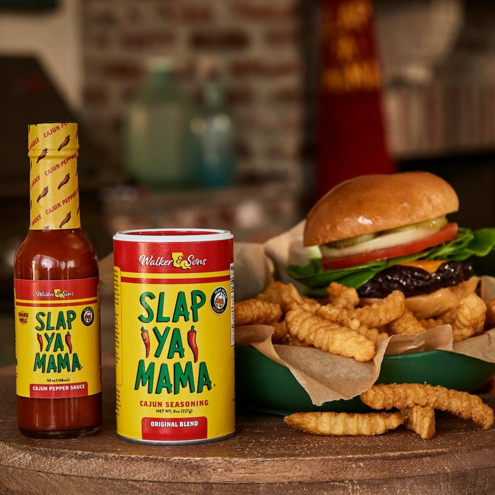 Cheeseburger and French fries on a plate next to Slap Ya Mama Cajun Seasoning and Cajun Pepper Sauce, with an outdoor kitchen in the background