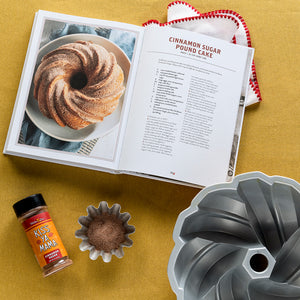 
                  
                    Load image into Gallery viewer, Open cookbook with cinnamon sugar pound cake recipe, bundt cake pan, and cinnamon spice on a yellow surface.
                  
                