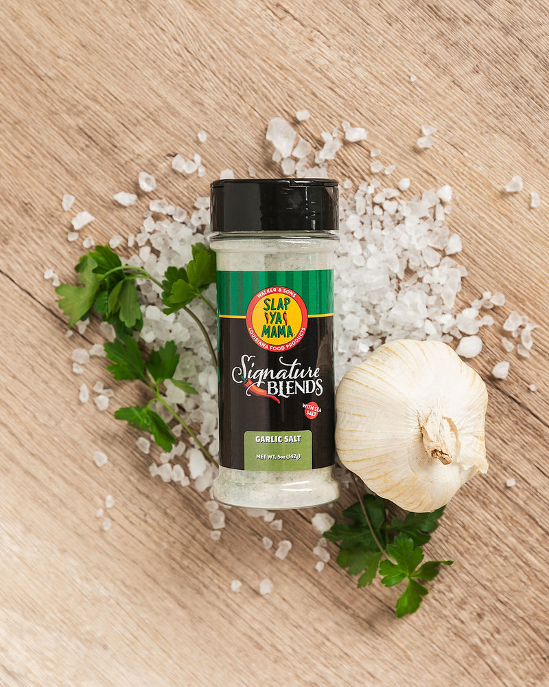 
                  
                    Load image into Gallery viewer, Lifestyle image of Slap Ya Mama Garlic Salt Signature Blend canister laying on sea salt, with head of garlic and fresh parsley scattered around it
                  
                