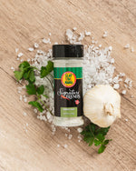 Lifestyle image of Slap Ya Mama Garlic Salt Signature Blend canister laying on sea salt, with head of garlic and fresh parsley scattered around it