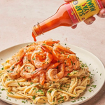 Image of shrimp Alfredo pasta with Slap Ya Mama Cajun Pepper Sauce being poured over it. The dish is garnished with parsley on a white plate.