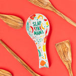 Slap Ya Mama white spoon rest with a green logo, featuring illustrated beignets and crawfish, styled with a red background and wooden spoons for a rustic, vibrant look.