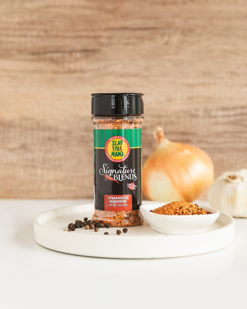 
                  
                    Load image into Gallery viewer, Slap Ya Mama Signature Blend Steakhouse Seasoning canister styled with fresh ingredients, including onion, garlic, and poured-out seasoning on a clean white countertop, creating a rustic and flavorful display
                  
                