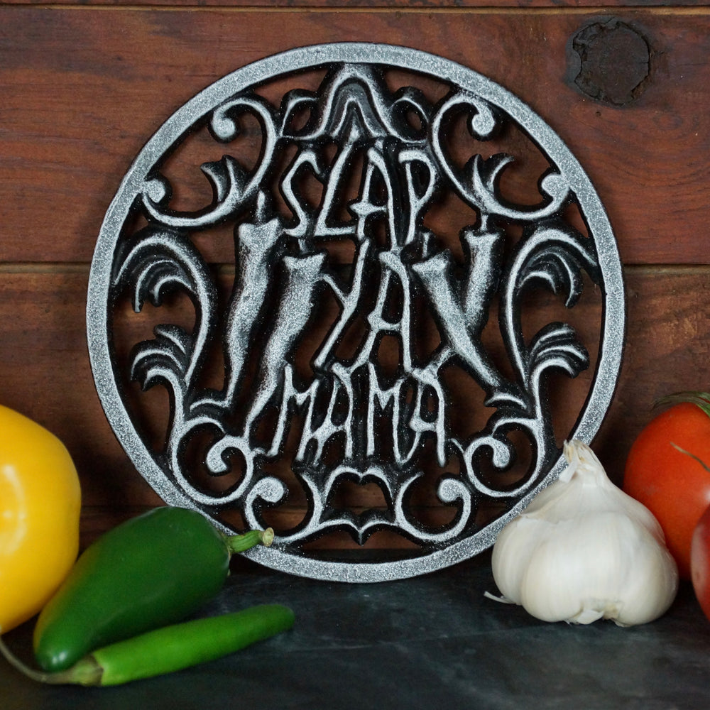 7-inch cast iron trivet with the Slap Ya Mama logo, placed on a wooden background with fresh vegetables surrounding it, creating a rustic and flavorful scene.