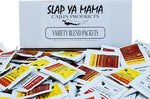 Case of 100 Slap Ya Mama seasoning packets, including 25 Original Blend Packets, 25 Hot Blend Packets, 25 White Pepper Blend Packets, and 25 Low Sodium Blend Packets. Displayed with a pile of seasoning packets in front of a white box featuring the Slap Ya Mama logo in black.